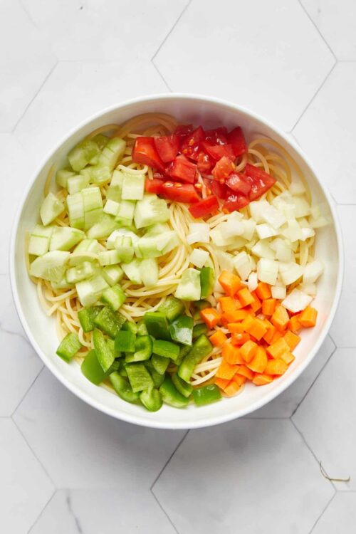 Colorful vegetable pasta salad with chopped cucumbers, tomatoes, carrots, and bell peppers in a white bowl.