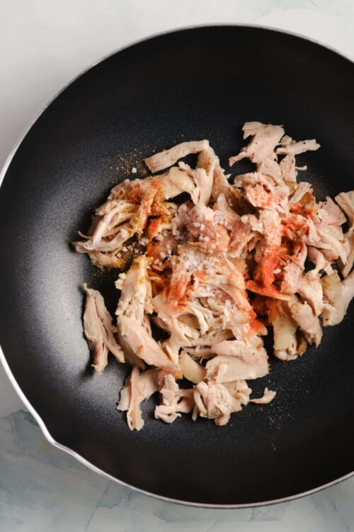 Shredded cooked chicken in a non-stick skillet with spices and seasoning for baking Bree recipes.