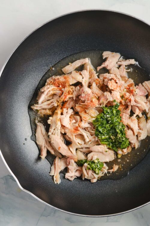 Shredded cooked chicken in a black skillet with chopped herbs and spices for recipe preparation.