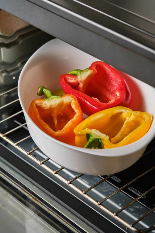 Colorful bell peppers roasting in a white bowl inside an oven.