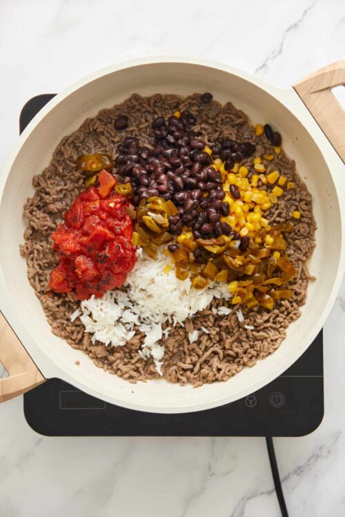 Ground beef and rice in a skillet with tomatoes, black beans, corn, and pickled jalapenos, ready for cooking or serving.