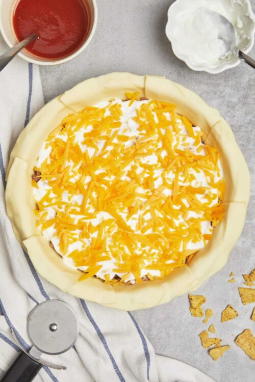 Shredded cheddar cheese and sour cream on unbaked cheese and beef taco pie.
