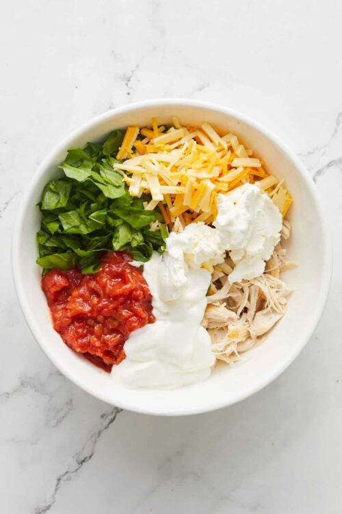 Shredded cheese, lettuce, tomatoes, chicken, sour cream, and salsa in a white bowl for Mexican casserole.