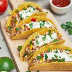 Freshly made Mexican tacos with seasoned meat, corn, and topped with sour cream and cilantro on a wooden serving board.