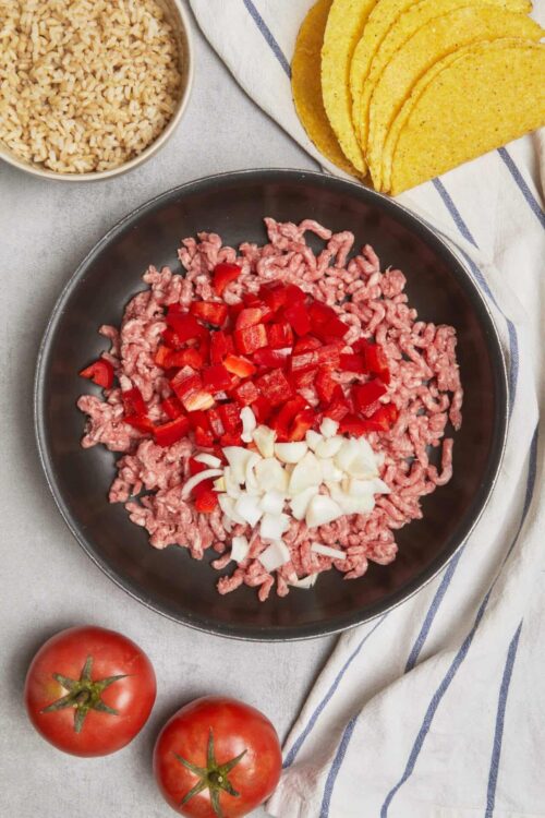 Ground beef with chopped onions and red bell peppers in a skillet, complemented by rice, tortillas, and fresh tomatoes.