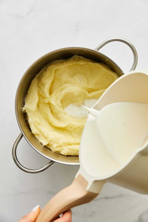 Creamy mashed potatoes being prepared with milk, butter, and a potato masher.