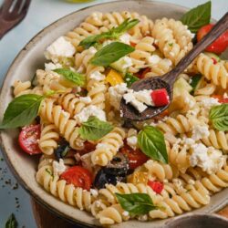 Fusilli pasta salad with cherry tomatoes, fresh basil, black olives, and feta cheese, served in a rustic bowl.