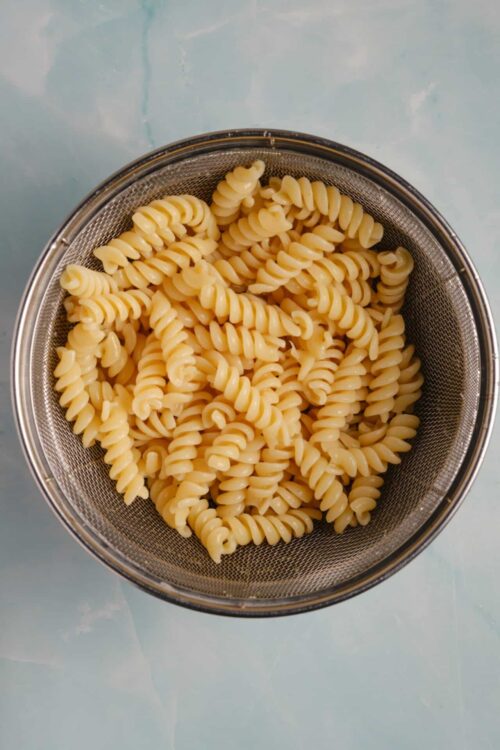 Cooked rotini pasta in a metal strainer on a light blue surface.