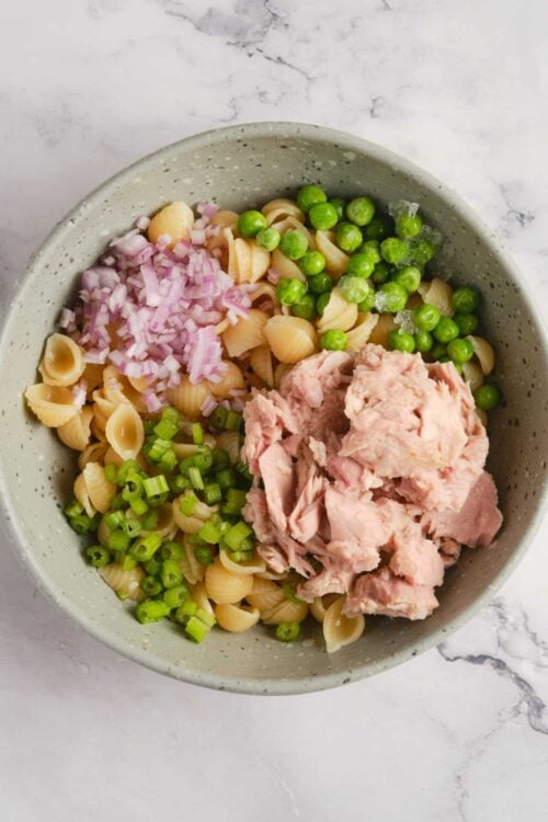 Cooked pasta with chopped red onions, frozen peas, chopped celery, and shredded cooked chicken in a speckled grey bowl.