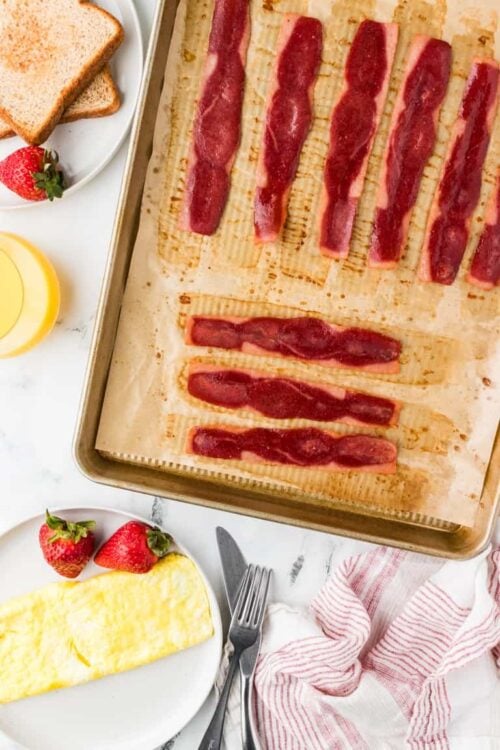 Baked bacon strips on baking sheet with breakfast staples and strawberries for a delicious morning meal.