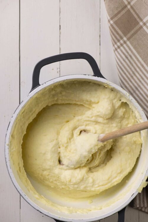 Creamy mashed potatoes in a white pot with a black handle, on a rustic white wooden surface.