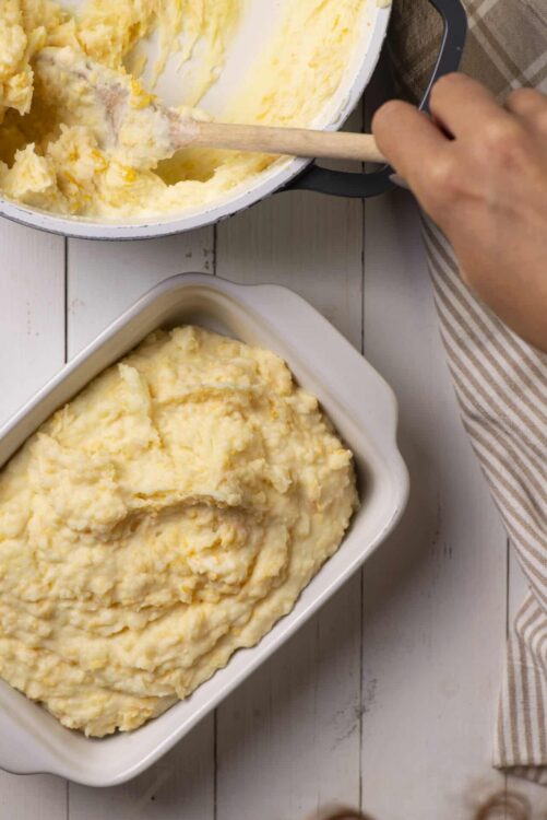 Creamy mashed potatoes in a baking dish with a hand mashing in a pot; homemade comfort food.