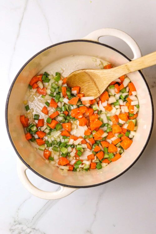 Chopped celery, carrots, and onions cooking in a white enamel pot with a wooden spoon.