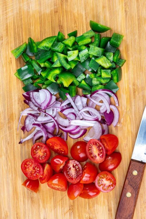 Chopped green bell peppers, red onions, and cherry tomatoes on wooden cutting board.