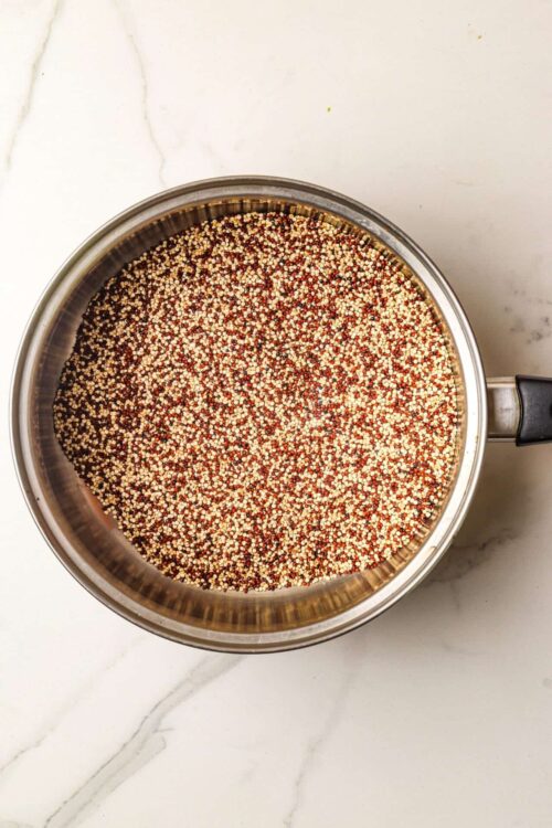 Colored quinoa seeds in a stainless steel pot on a white marble surface.