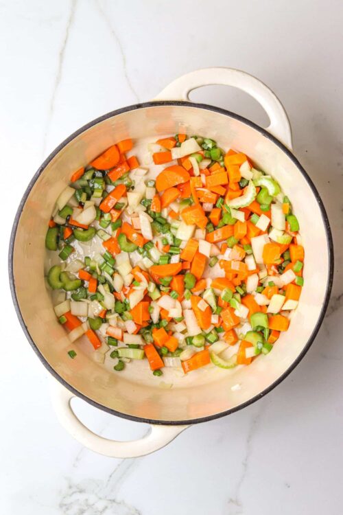 Diced onions, carrots, celery cooking in a white cast-iron skillet for vegetable sautéing.