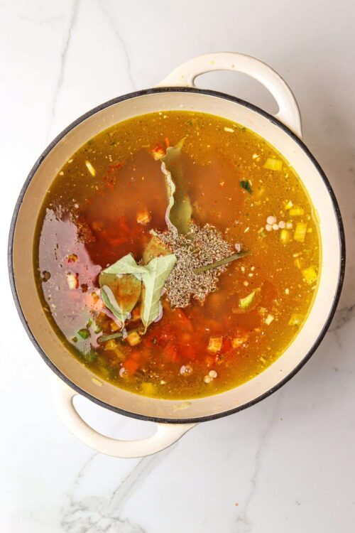 Aromatic homemade vegetable chicken soup with herbs in a white pot on marble countertop.