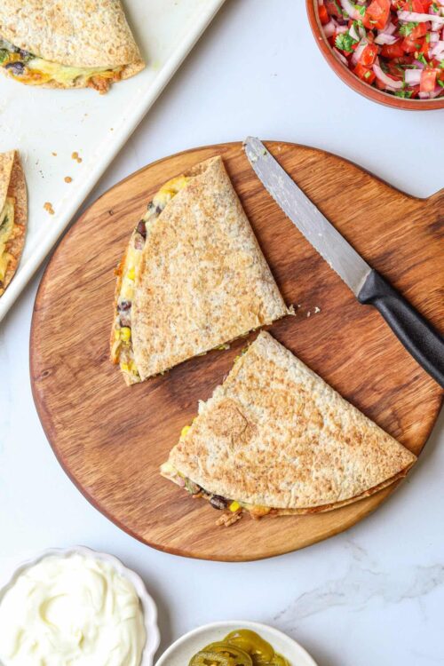 Crispy quesadilla on wooden cutting board with black handled knife, fresh salsa, sour cream, and jalapenos in background.