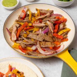 Juicy grilled beef fajitas with colorful peppers and onions on a yellow cast iron skillet.