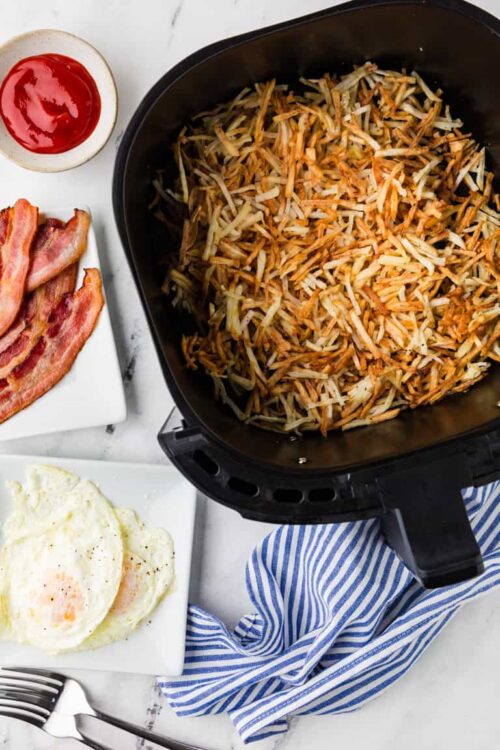 Crunchy hash browns cooking in an air fryer with crispy bacon and fried eggs on a white plate.