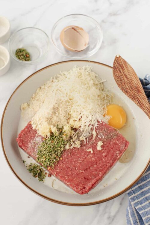 Fresh ground beef and ingredients for meatloaf with shredded cheese, eggs, herbs, and spices on a white marble countertop.