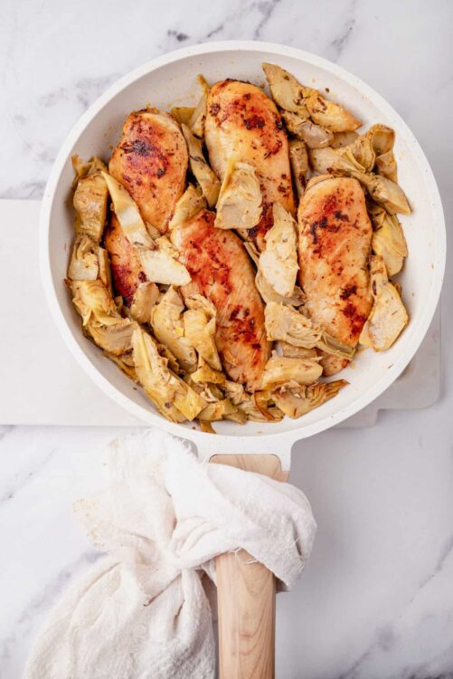 Roasted chicken breasts with artichokes in a white casserole dish on marble surface.