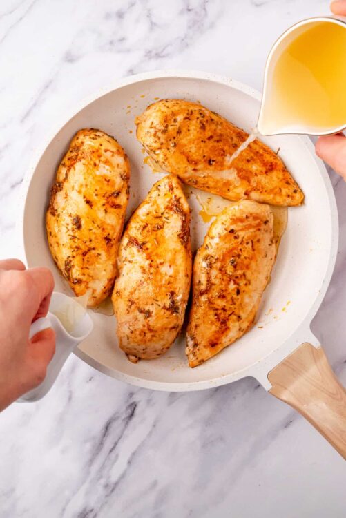Juicy baked chicken breasts being drizzled with lemon sauce in a white skillet.
