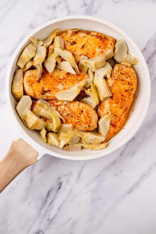 Juicy baked chicken breasts with artichoke hearts in a white skillet close-up.