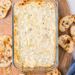 Creamy cheesy baked dip in a glass dish with toasted baguette slices on a wooden board.
