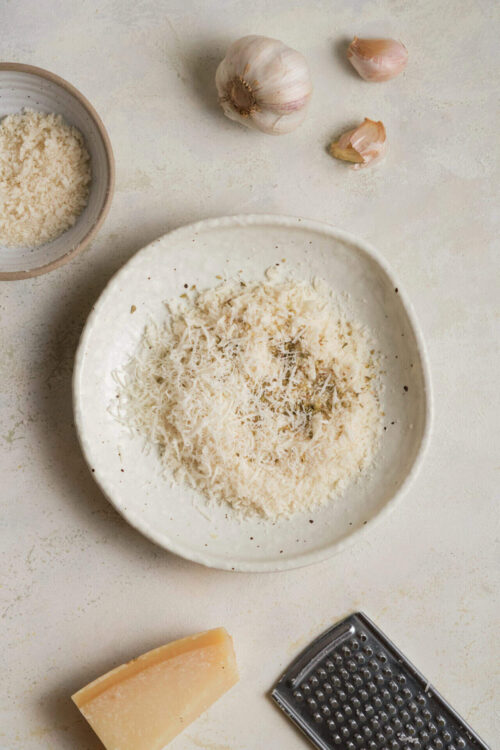 Shredded cheese, garlic, and parmesan cheese on a white plate with garlic bulbs and cheese grater.