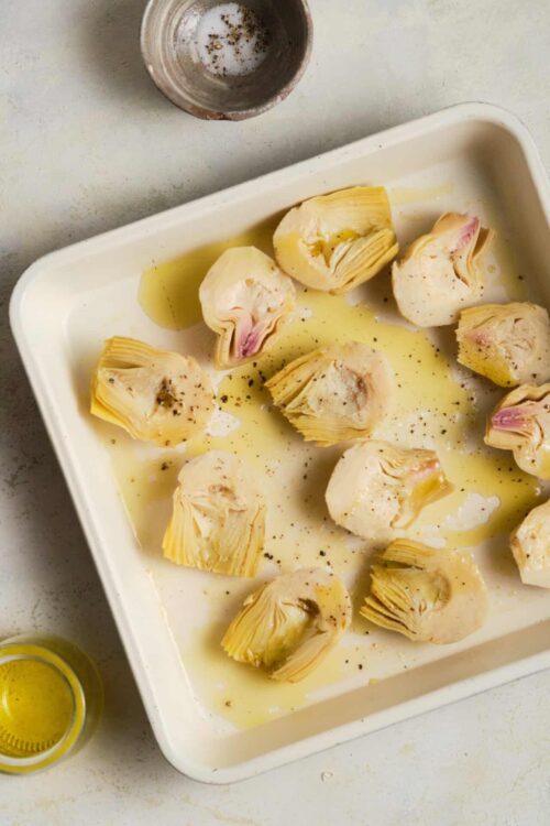 Artichoke hearts in olive oil with salt and pepper in a white baking dish.
