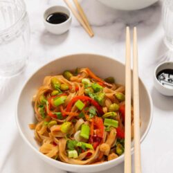 Stir-fried vegetable and noodle dish served in a white bowl with chopsticks on a white table.