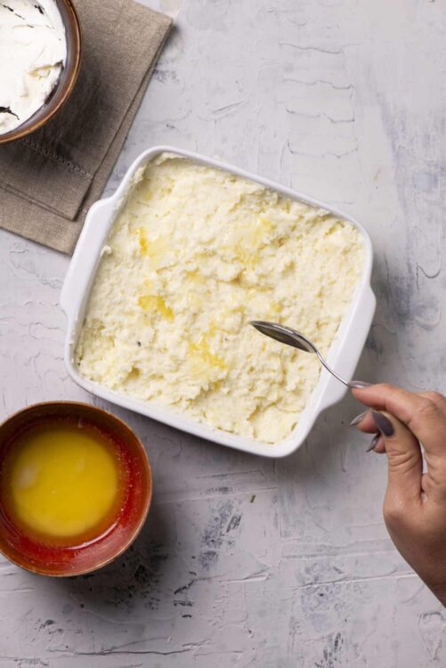 Creamy mashed potatoes in a white baking dish with a hand holding a spoon.