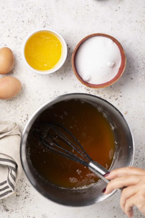 Sweet caramel sauce being whisked in a stainless steel pot on a light countertop with baking ingredients.