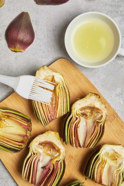 Artichokes being brushed with olive oil on a wooden cutting board with a small bowl of olive oil, fresh artichoke, and a basting brush, ready for roasting or grilling.
