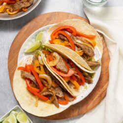 Sizzling beef fajitas with colorful peppers on a white plate, served with lime wedges, guacamole, and fresh water, on a rustic wooden board.
