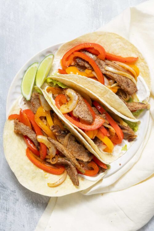 Grilled steak fajitas in soft tortillas with peppers and onions, lime wedges on white plate.