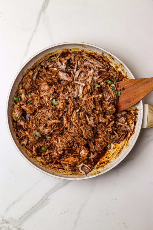 Shredded beef brisket in a skillet with sauce, garnished with herbs, and a wooden spatula inside.