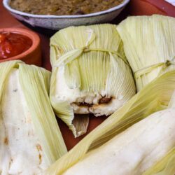 Fresh homemade tamales with corn husks on a rustic plate, served with salsa and Mexican side dishes.