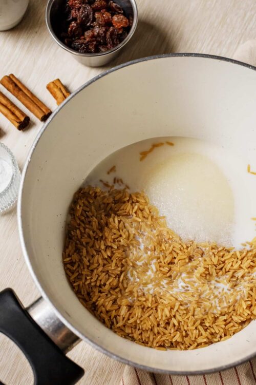 Cooked brown rice in a pot with milk and cinnamon sticks on a wooden table.