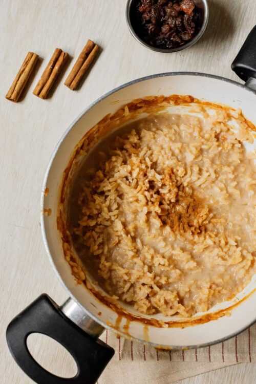 Creamy rice pudding cooking in a white pot with cinnamon and raisins on a wooden surface.