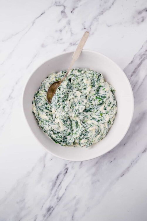Creamed spinach with cheese in a white bowl on marble surface.