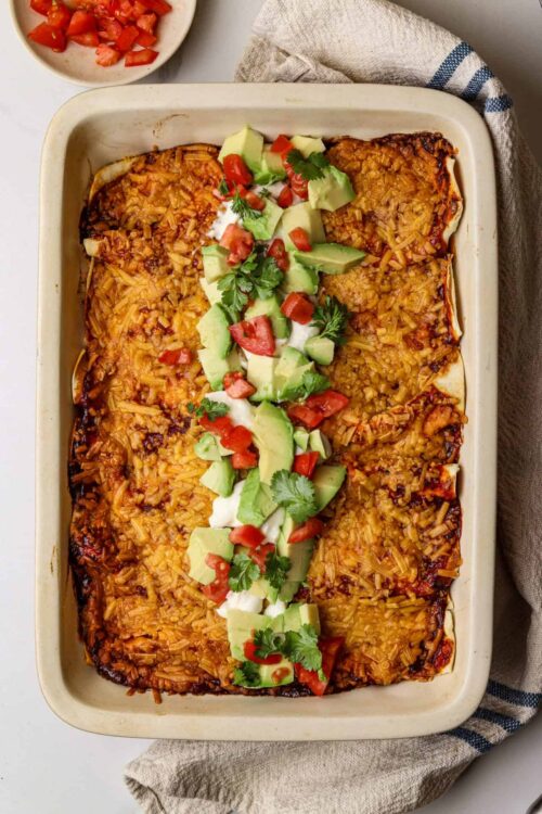 Baked enchiladas in a baking dish topped with diced tomatoes, avocado, cilantro, and sour cream for a delicious Mexican dinner.