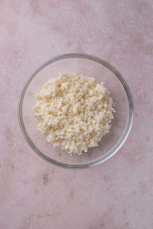 Crumbled white cheese in a glass bowl on pink textured background.