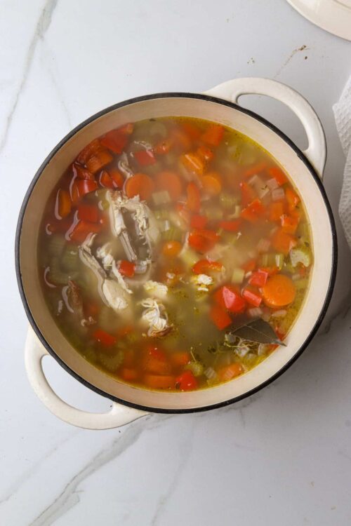 Bouillon with vegetables and chicken for homemade soup, in a white enamel pot on a marble countertop.