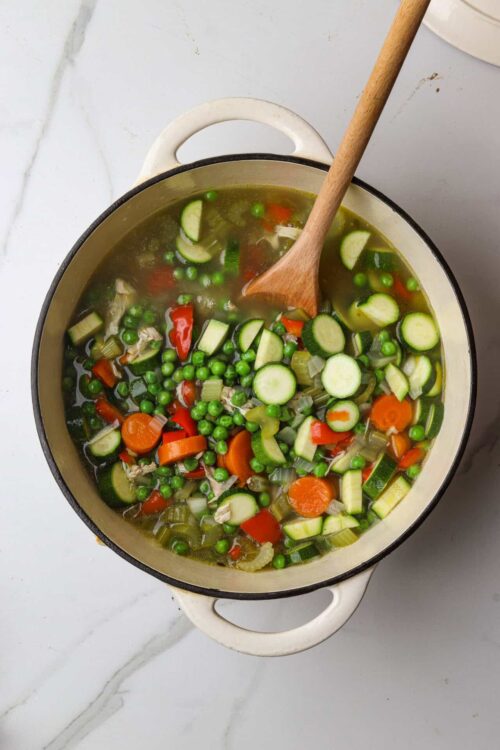 Fresh vegetable soup with peas, zucchini, carrots, and celery in a white pot.