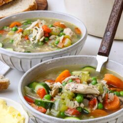 Homemade chicken vegetable soup served in rustic bowls with bread slices and mashed potatoes on a white table.