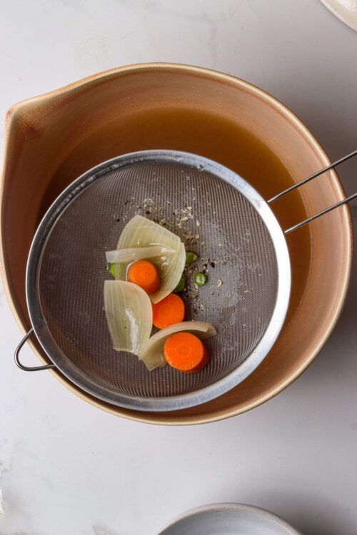 Boiled onions, carrots, and garlic in a colander over a pot, preparing vegetables for cooking.