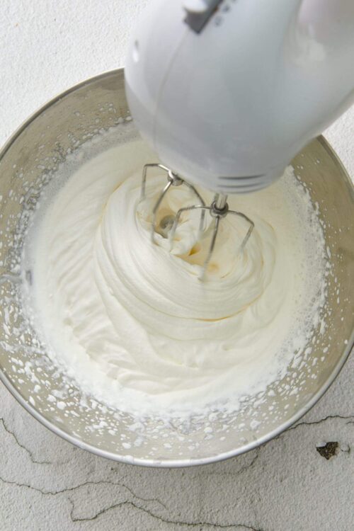 Fluffy whipped cream being mixed with a hand mixer in a stainless steel bowl.
