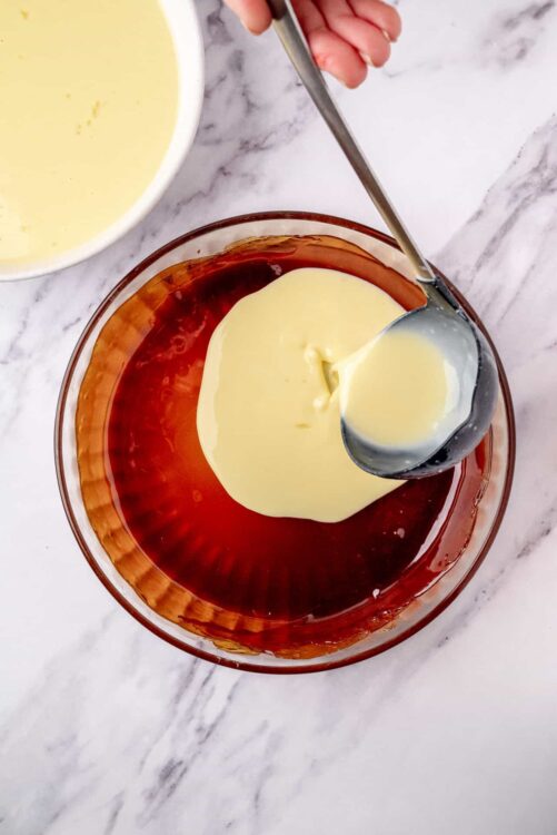 Cream being poured into a bowl of red gelatin for a dessert recipe.